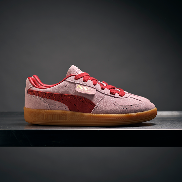 Rosa og røde Puma Palermo joggesko med gummisåle står på en metallhylle mot en mørk bakgrunn.