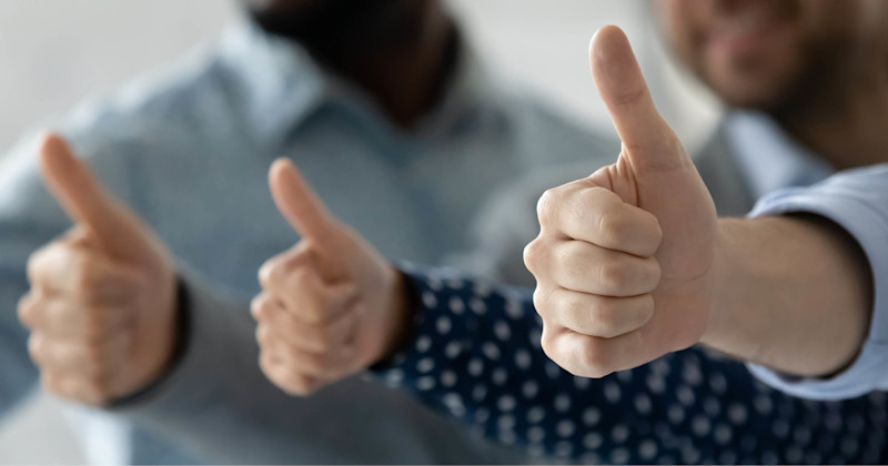 Tre personer gir tommel opp-tegn med fokus på hendene i forgrunnen i et lyst rom.