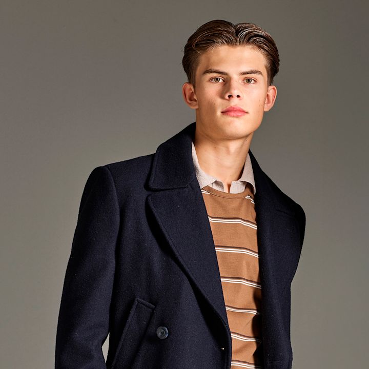 Portrett mot nøytral bakgrunn av person i mørkeblå frakk over brun stripete genser og lys skjorte.