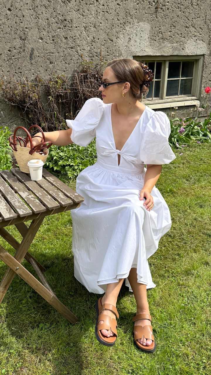 Kvinne i hvit kjole med puffermede ermer og brune sandaler sitter på gresset ved et trebord med en kaffekopp og veske i en hage med blomster og en steinvegg med vindu i bakgrunnen.