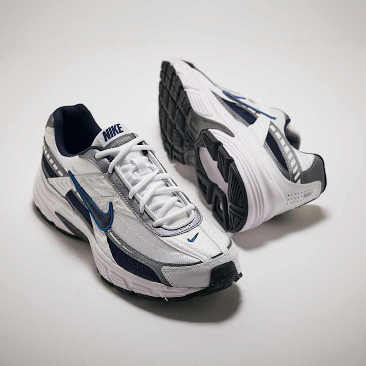 Hvite og grå Nike-løpesko med blå detaljer plassert på hvit bakgrunn, en sko viser overdelen og den andre sålen.