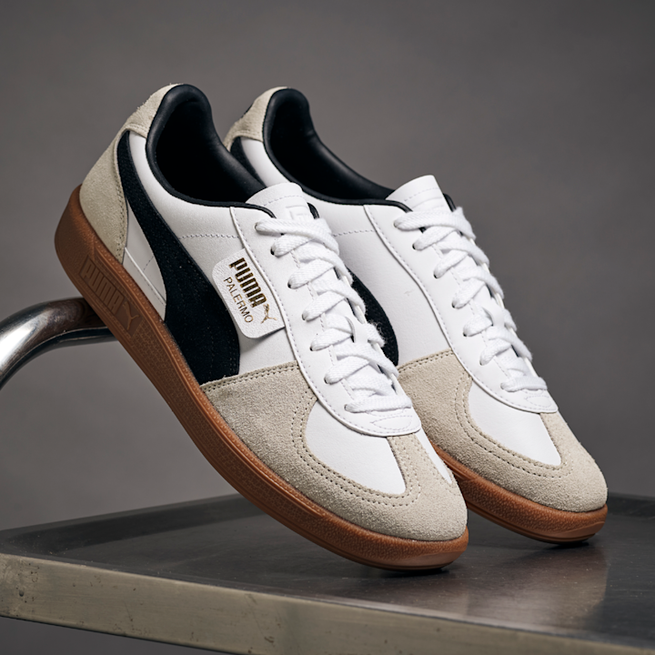 Hvite og beige Puma Palermo joggesko med svarte detaljer og brune såler plassert på en metallstol i et grått studio.