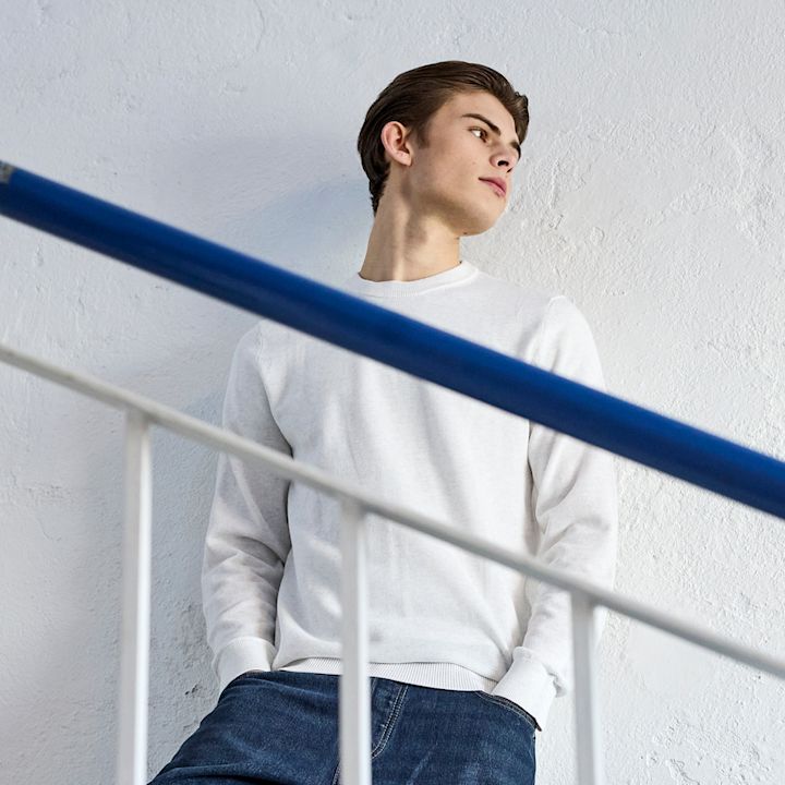 Ung mann i hvit genser og mørke blå jeans, lener seg mot en hvit vegg ved et blått rekkverk.