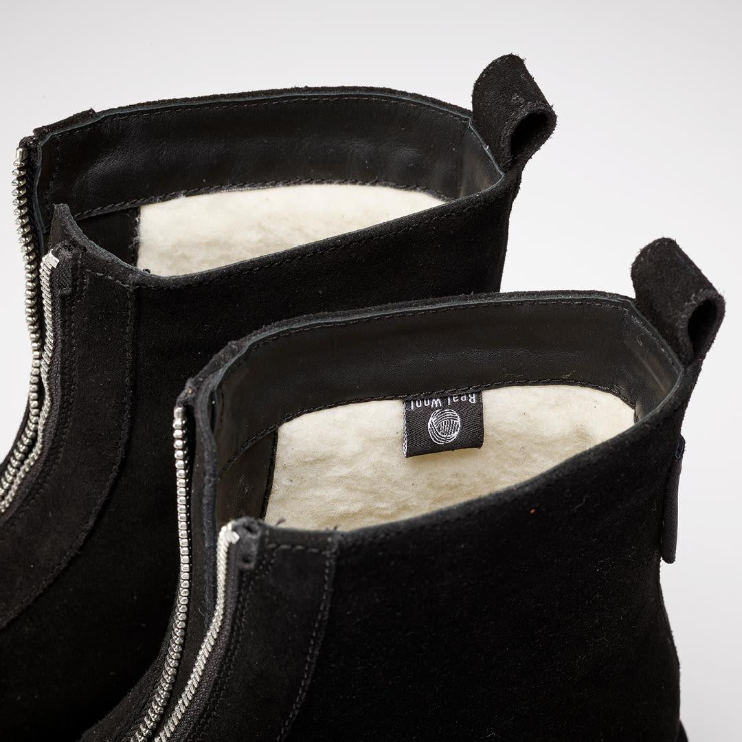 Svarte ankelstøvletter i semsket skinn med glidelås foran, hvitt ullfor og en merkelapp synlig på innsiden.