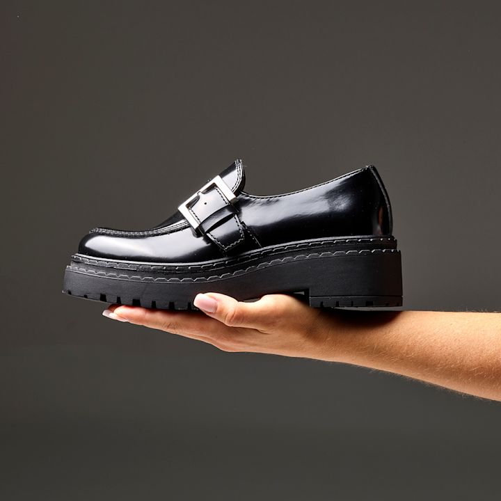 Svart, blank loafer med spenne holdt i en hånd. Skoen har kraftig, chunky såle og moderne, robust design.