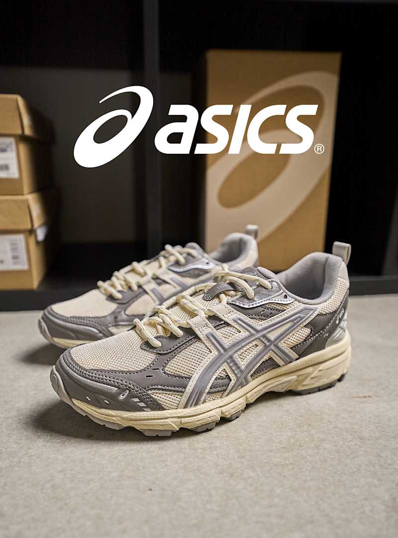 Asics løpesko i beige og grå med robust såle og retro stil, plassert foran Asics-logo og esker.