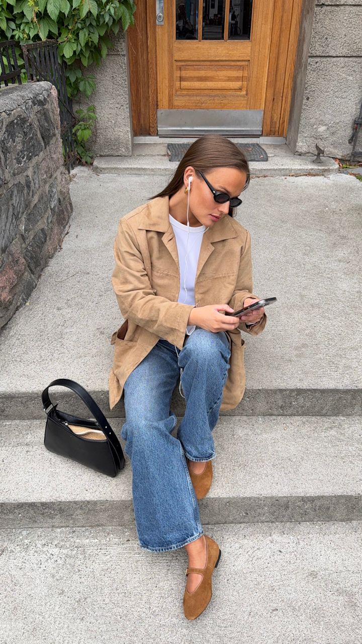 Kvinne med solbriller og beige jakke sitter på betongtrapp utenfor en bygning, bruker ørepropper og ser på mobilen med svart veske ved siden av seg.