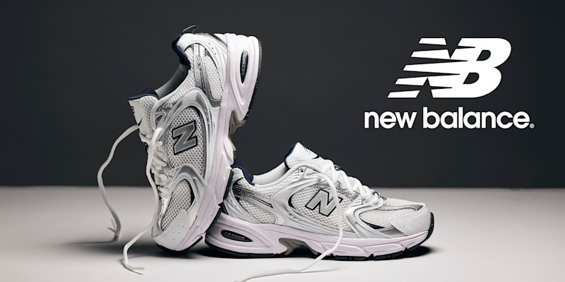 Hvite New Balance sneakers med mesh-overflate og sølv og marineblå detaljer, plassert ved siden av merkets logo på mørk bakgrunn.