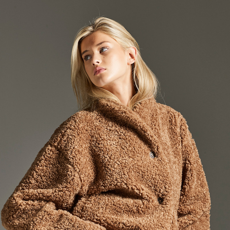 Person med lyst hår iført en lysebrun, fluffy teddyjakke mot en grå bakgrunn.