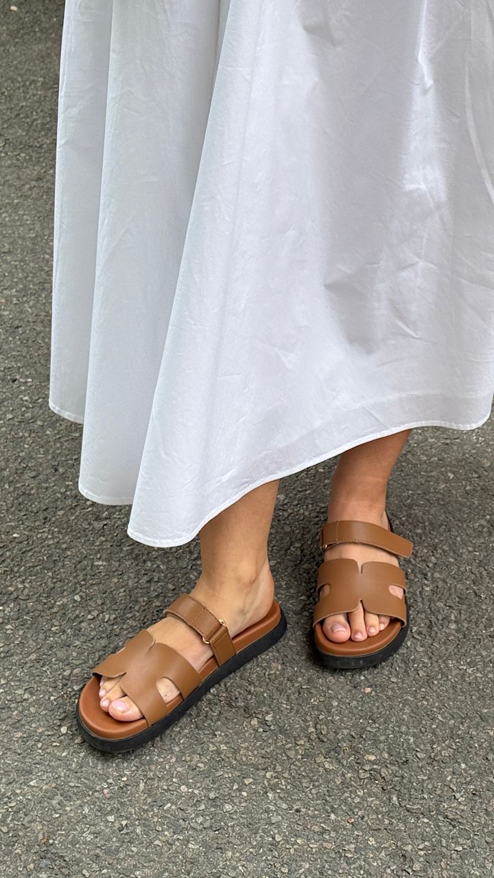 Brune sandaler med svart såle på føtter med hvit lang kjole som står på asfalt.